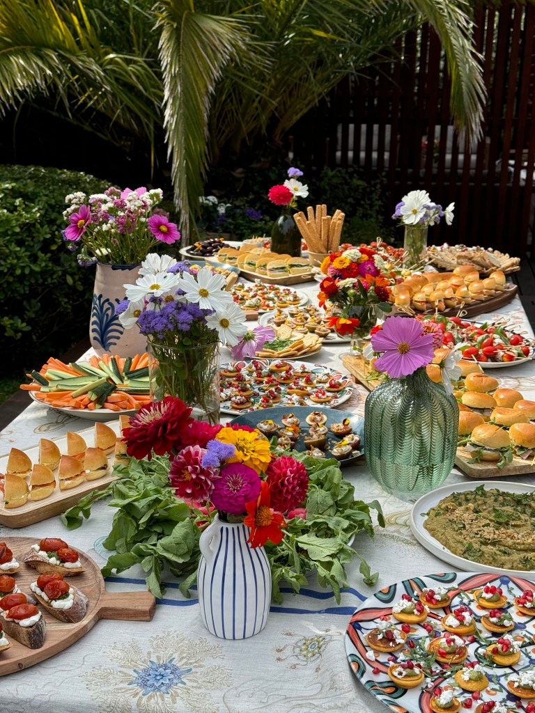 A vibrant outdoor table filled with an assortment of colorful appetizers, finger foods, and decorative flower arrangements in vases, set against a backdrop of lush greenery.